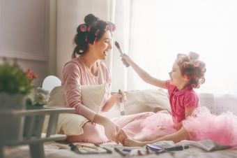 Mom sitting on floor with curlers in her hair while young daughter brushes makeup on her face