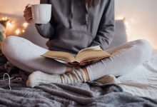 Cozy woman sitting in bed, mug in hand, reading a book.