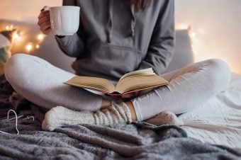 Cozy woman sitting in bed, mug in hand, reading a book.