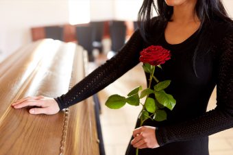 Woman at funeral wearing black dress standing by coffin holding a red rose
