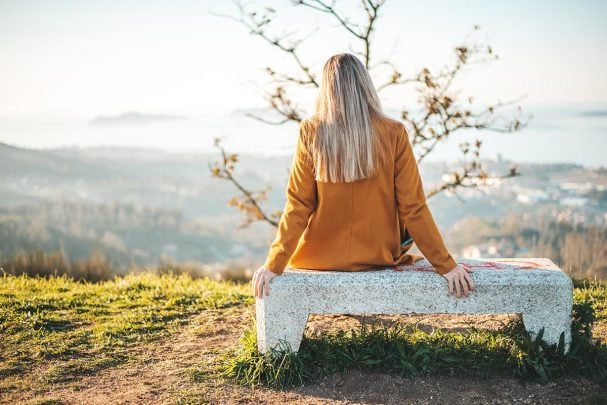 Woman in yellow coat sitting on a bench outside looking out at a nature view